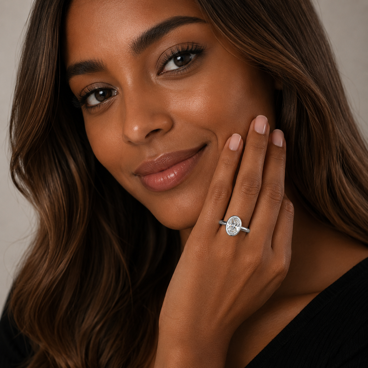 A close-up lifestyle shot of a woman with long brown hair wearing a silver ring with a round-cut clear gemstone on her finger, posing with her hand near her face against a neutral gray background.