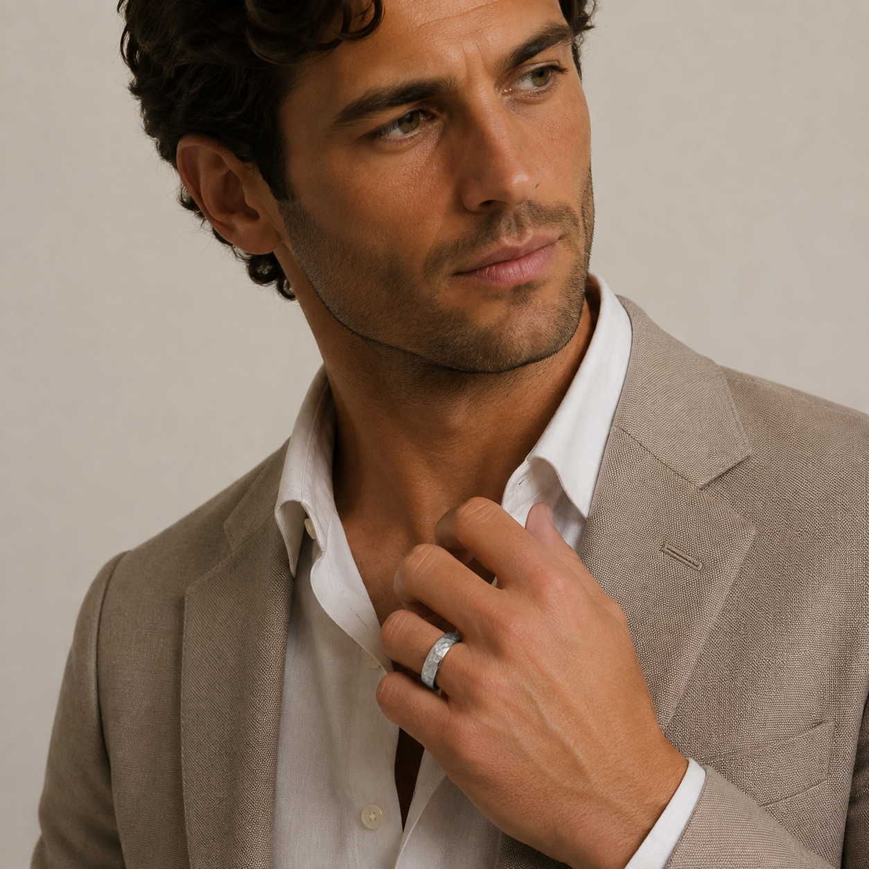 A close-up of a man wearing a light beige blazer over a white dress shirt, adjusting his collar while wearing a silver band ring on his finger, set against a neutral beige background.