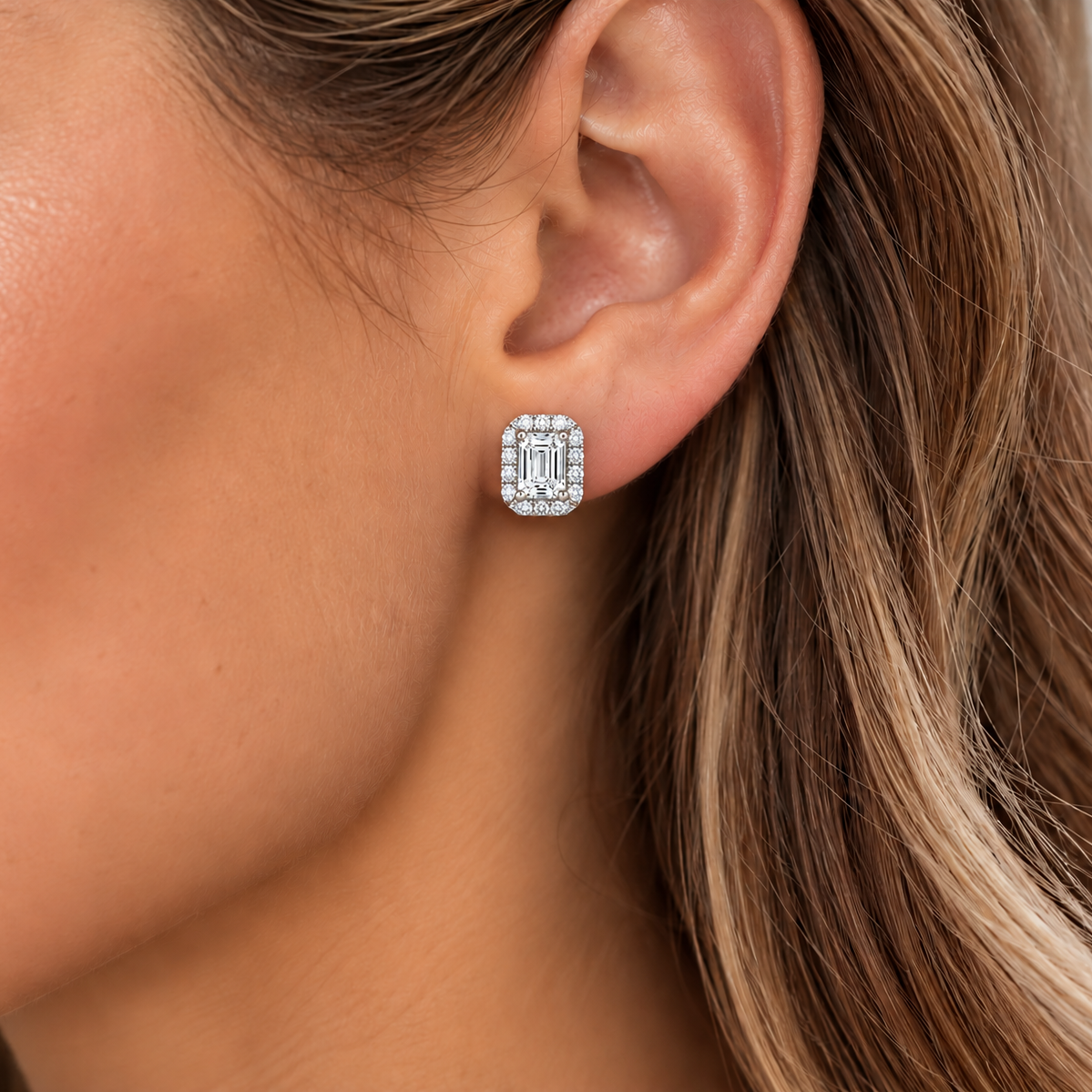 A close-up of a woman's ear wearing a small rectangular stud earring with a silver-toned border and a central baguette-cut clear gemstone.