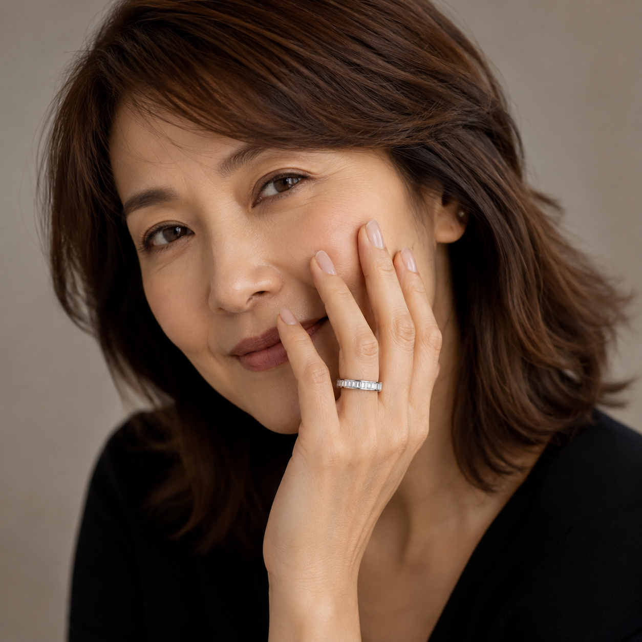 A close-up portrait of a woman with brown hair wearing a thin silver band ring with small clear stones on her finger, set against a soft beige background.