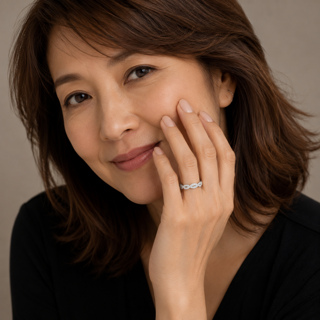 A close-up portrait of a woman with brown hair wearing a delicate silver ring with small clear stones on her finger, set against a soft beige background.
