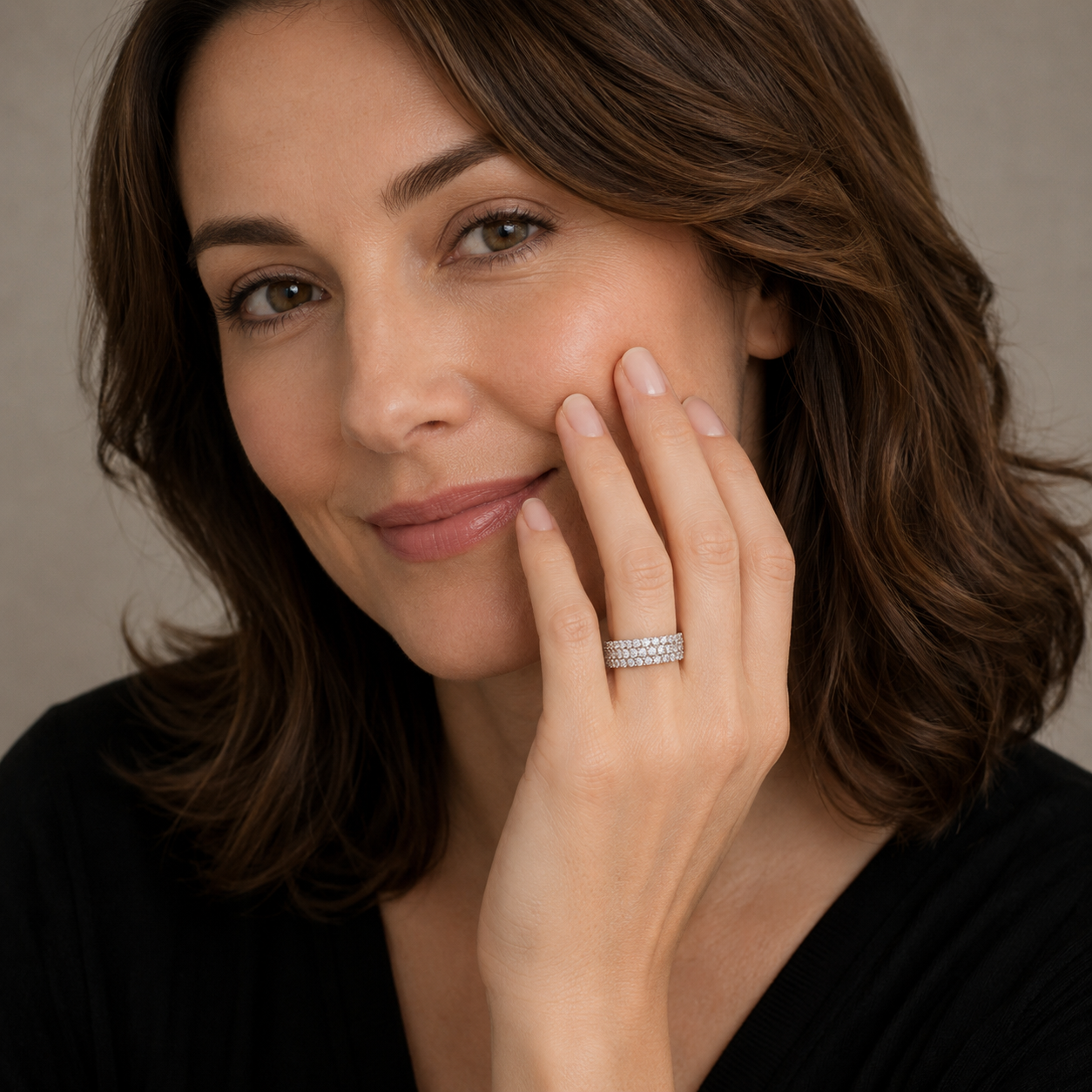 A close-up portrait of a woman with brown hair wearing a silver band ring set with small clear stones, touching her cheek. She is wearing a black top against a neutral beige background.
