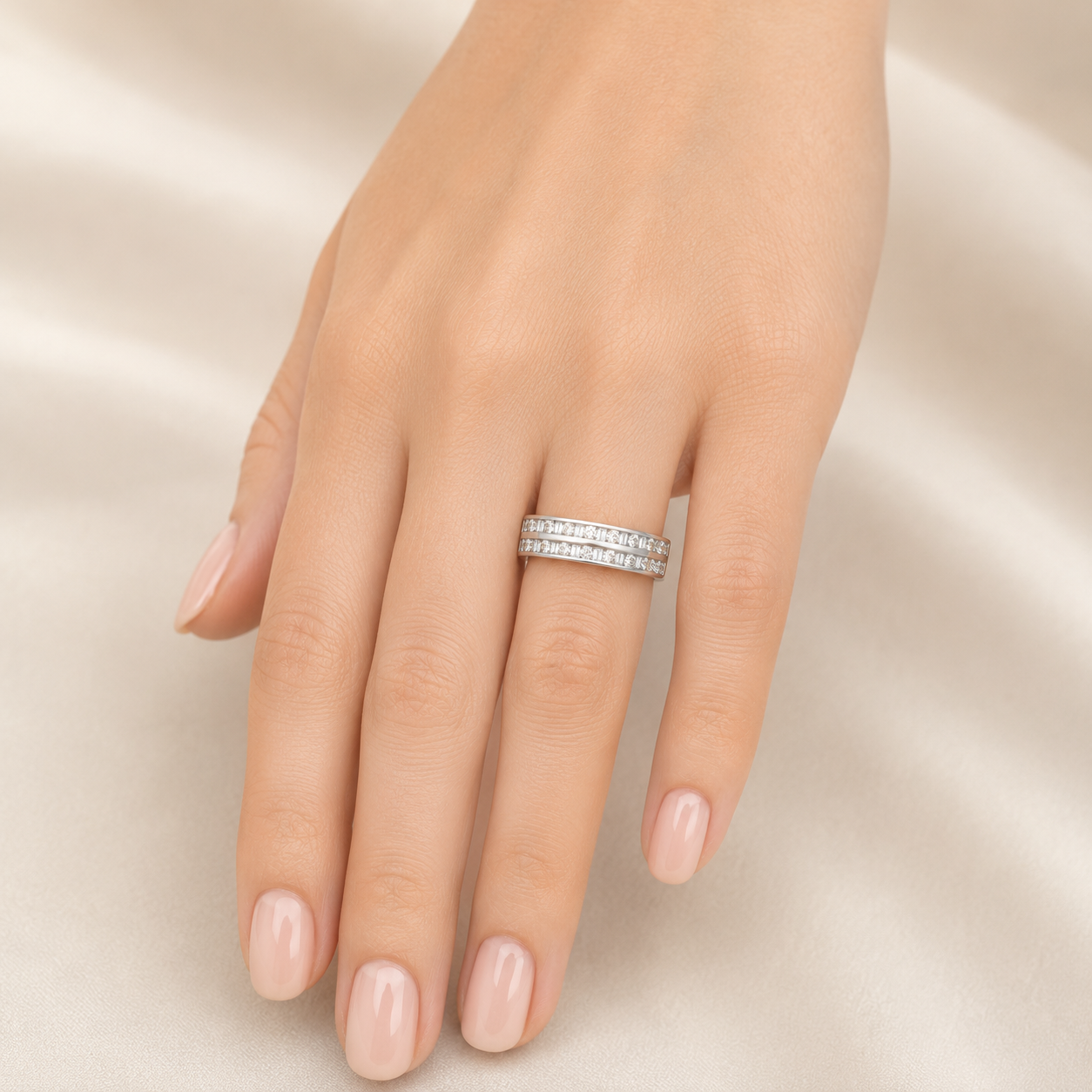 A close-up of a hand wearing a slim silver band ring set with small clear stones, photographed against a soft cream satin background.
