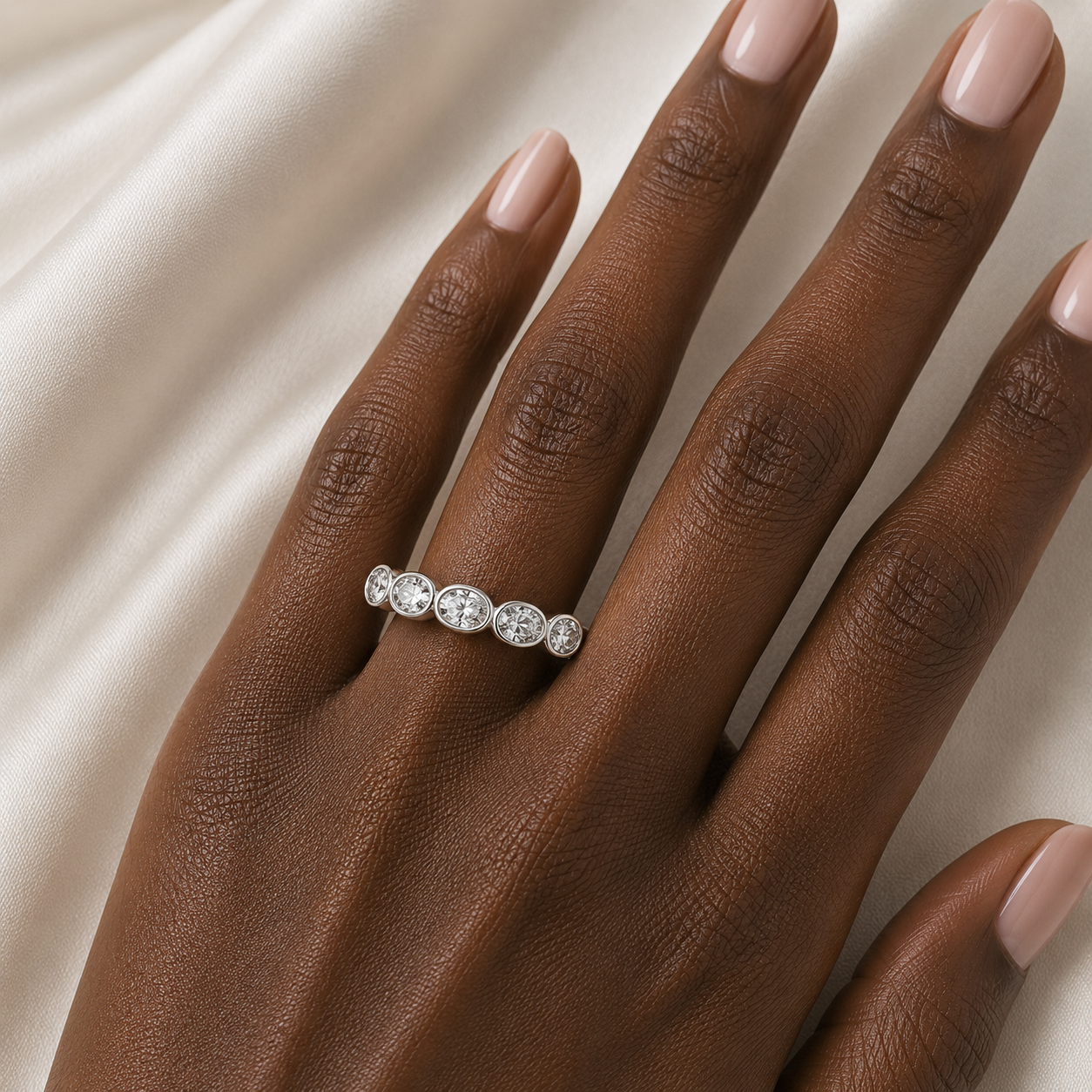 A close-up of a hand with pale pink manicured nails wearing a silver band ring set with five round clear gemstones, resting against a draped cream fabric background.