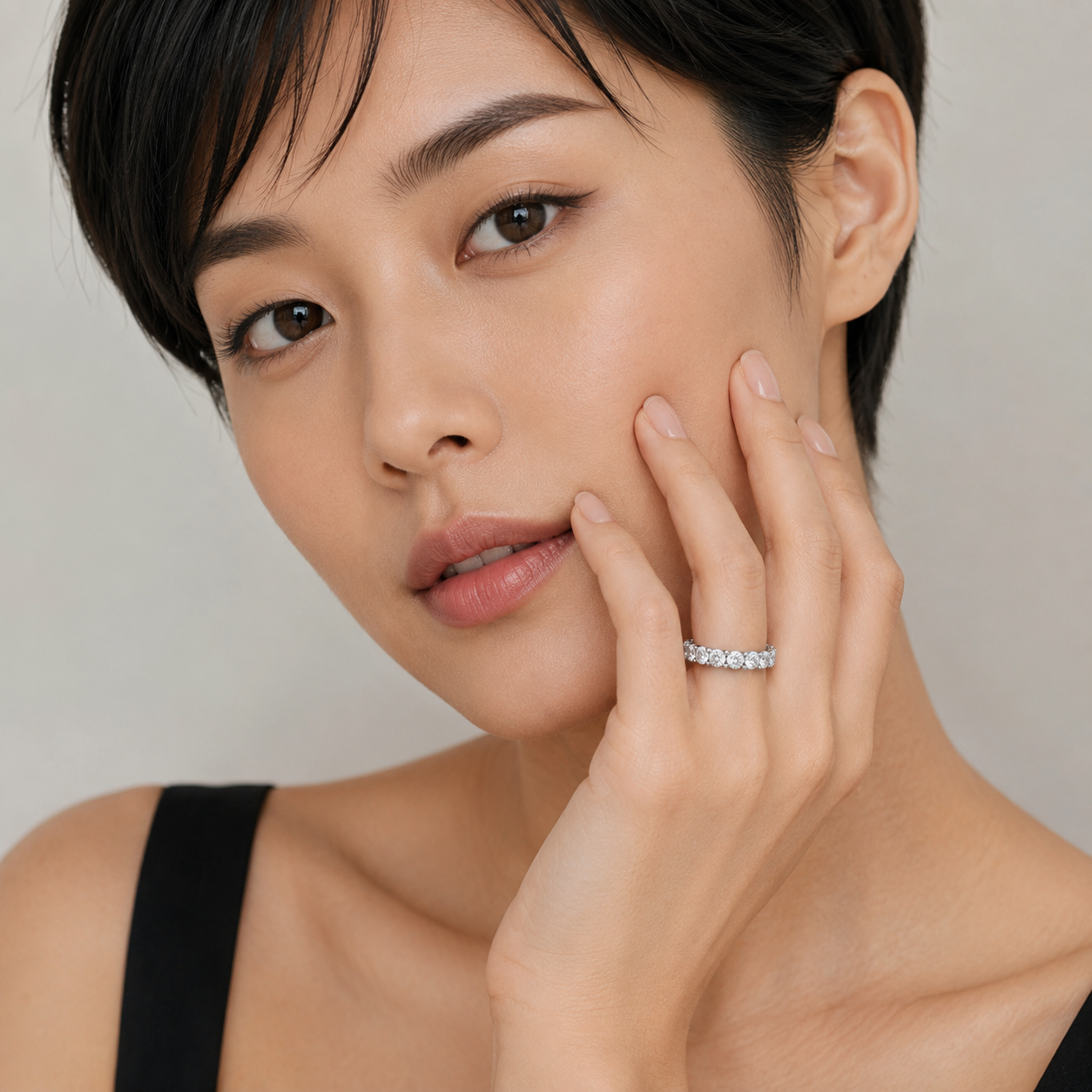 A close-up portrait of a woman with short dark hair wearing a thin silver band ring set with small clear stones, touching her cheek against a soft light gray background.