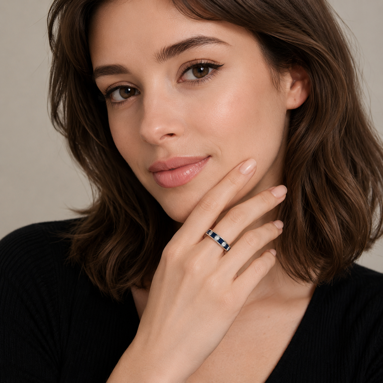 A close-up of a woman with brown hair wearing a slim silver band ring with small dark blue square accents, resting her chin on her hand against a soft beige background.