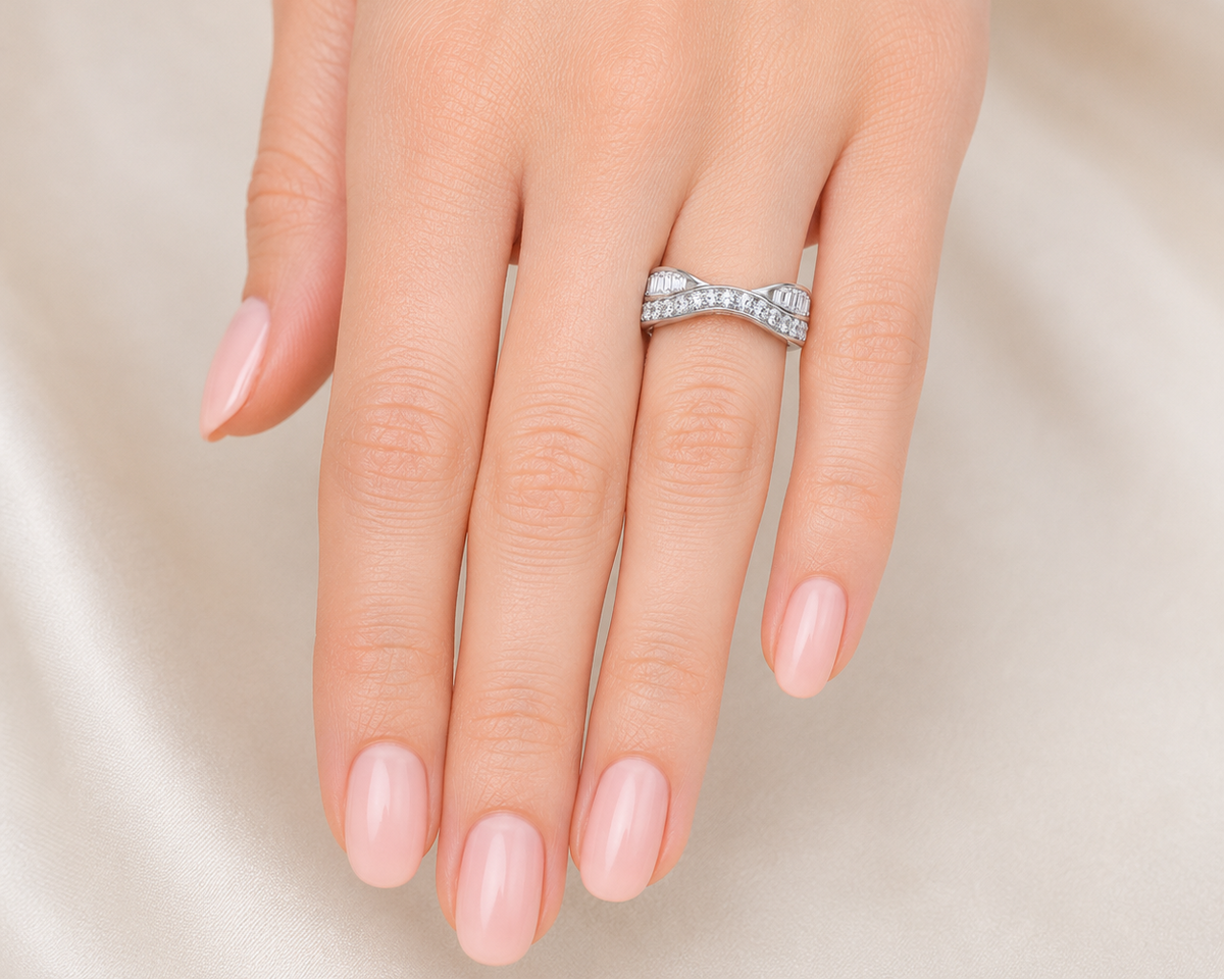 A close-up of a hand with pale pink manicured nails wearing a silver ring set with small clear stones in a bow-like twisted design, against a soft cream satin background.