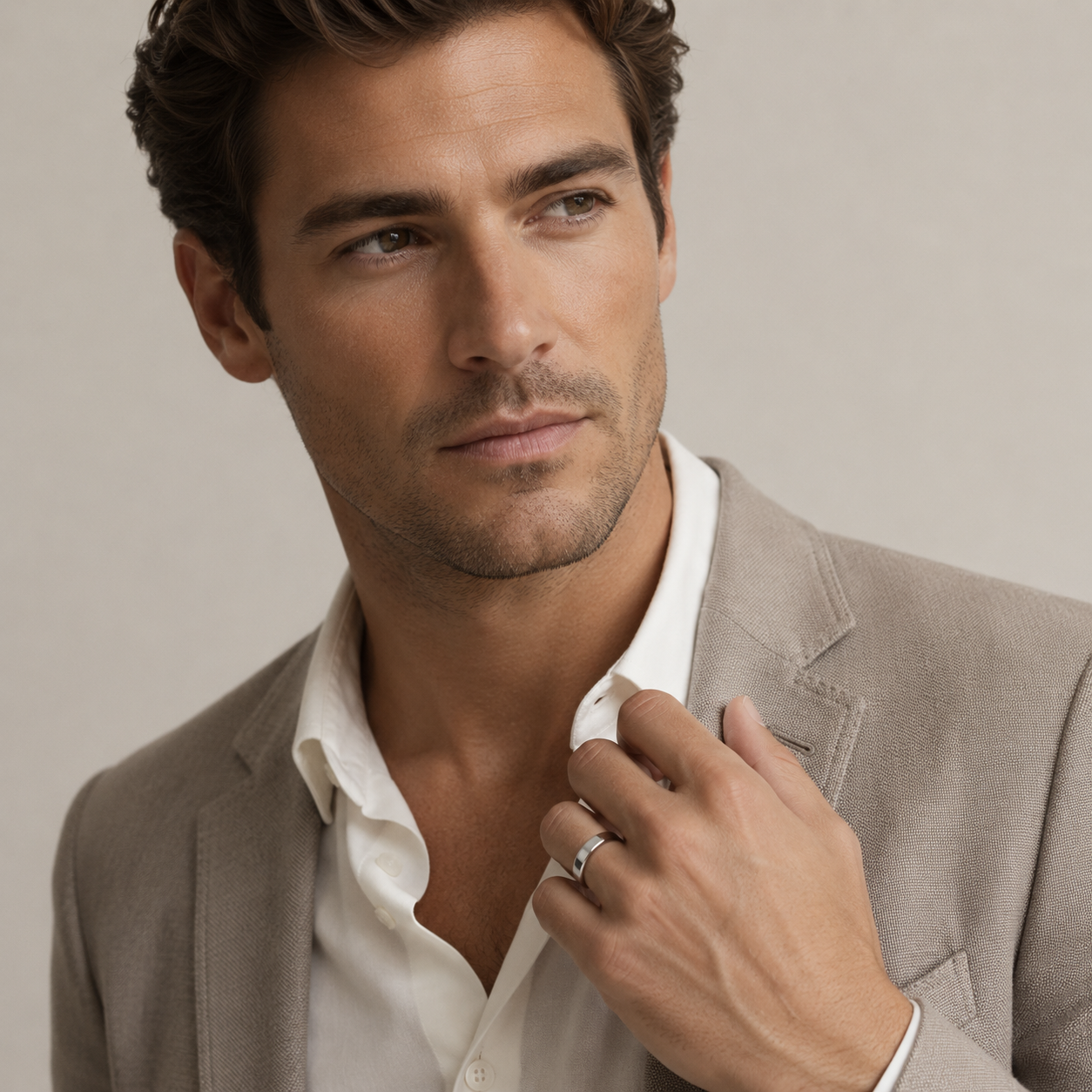 A close-up portrait of a man wearing a light beige blazer over an open white shirt, with one hand adjusting the collar and the other holding a ring. The background is a soft, neutral beige studio backdrop.