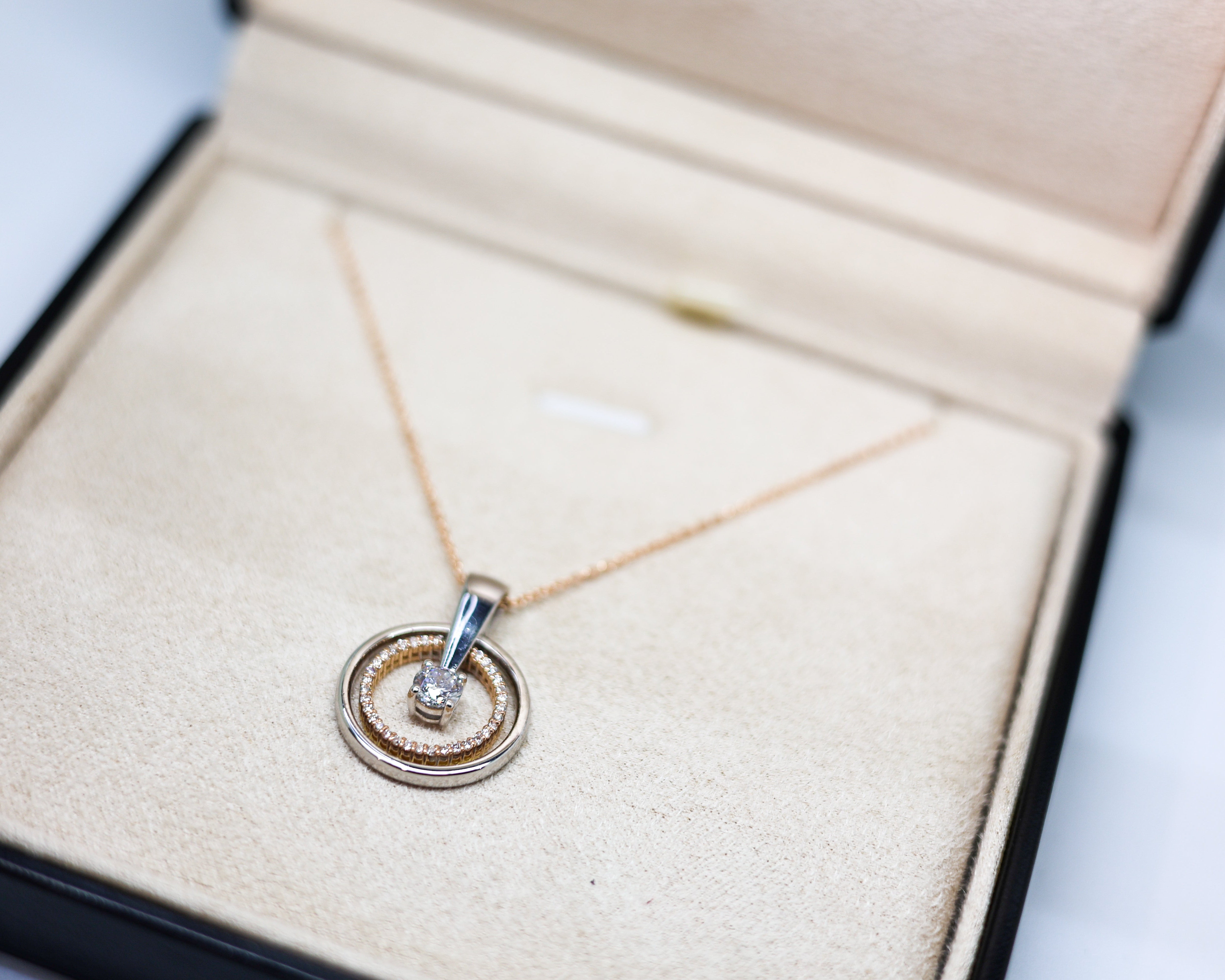 A delicate gold chain necklace with a double-ring pendant, one of which is set with a small clear gemstone, displayed inside an open beige jewelry box.