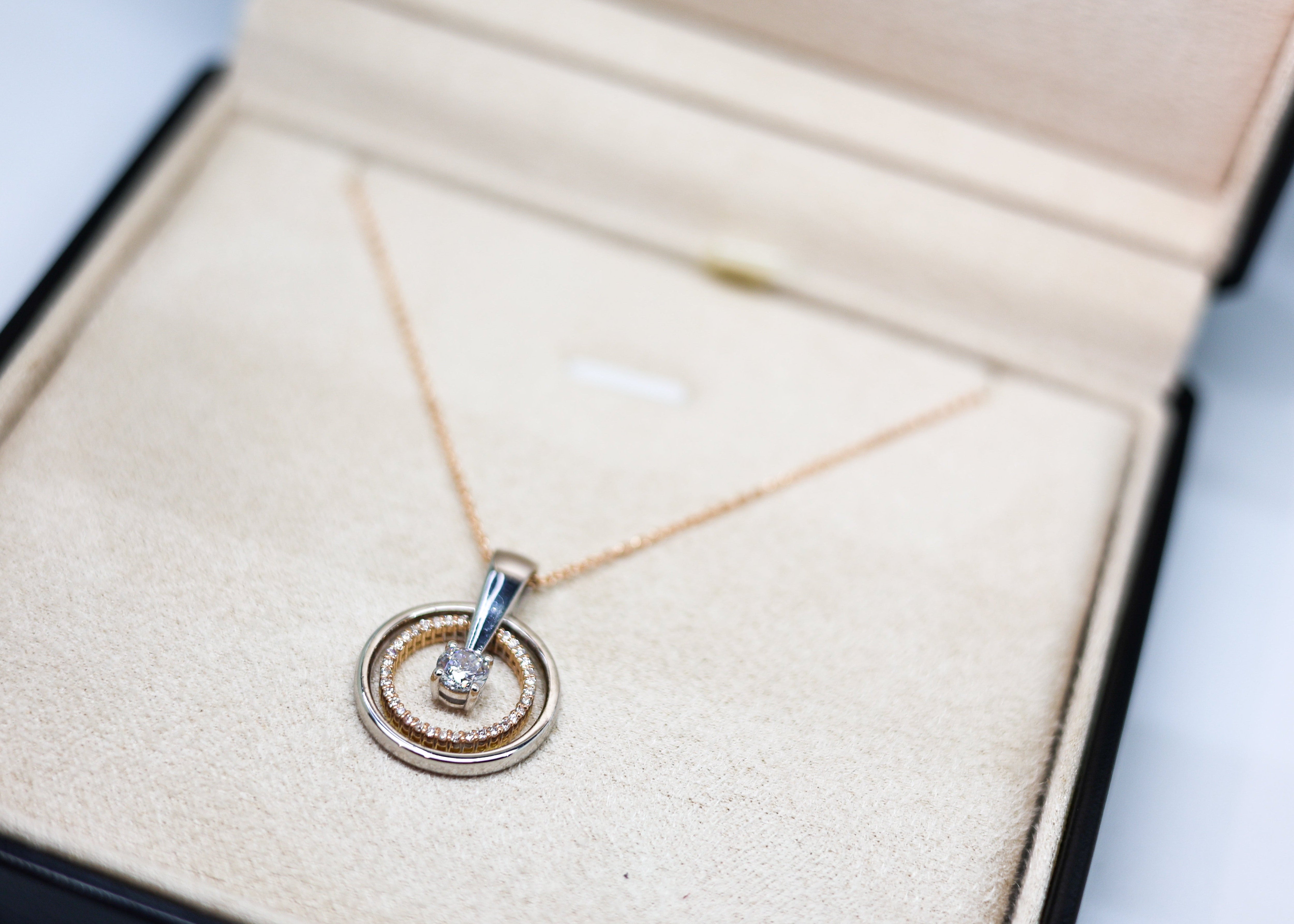 A delicate gold chain necklace with a double-ring pendant, one of which is set with a small clear gemstone, displayed inside an open beige jewelry box.