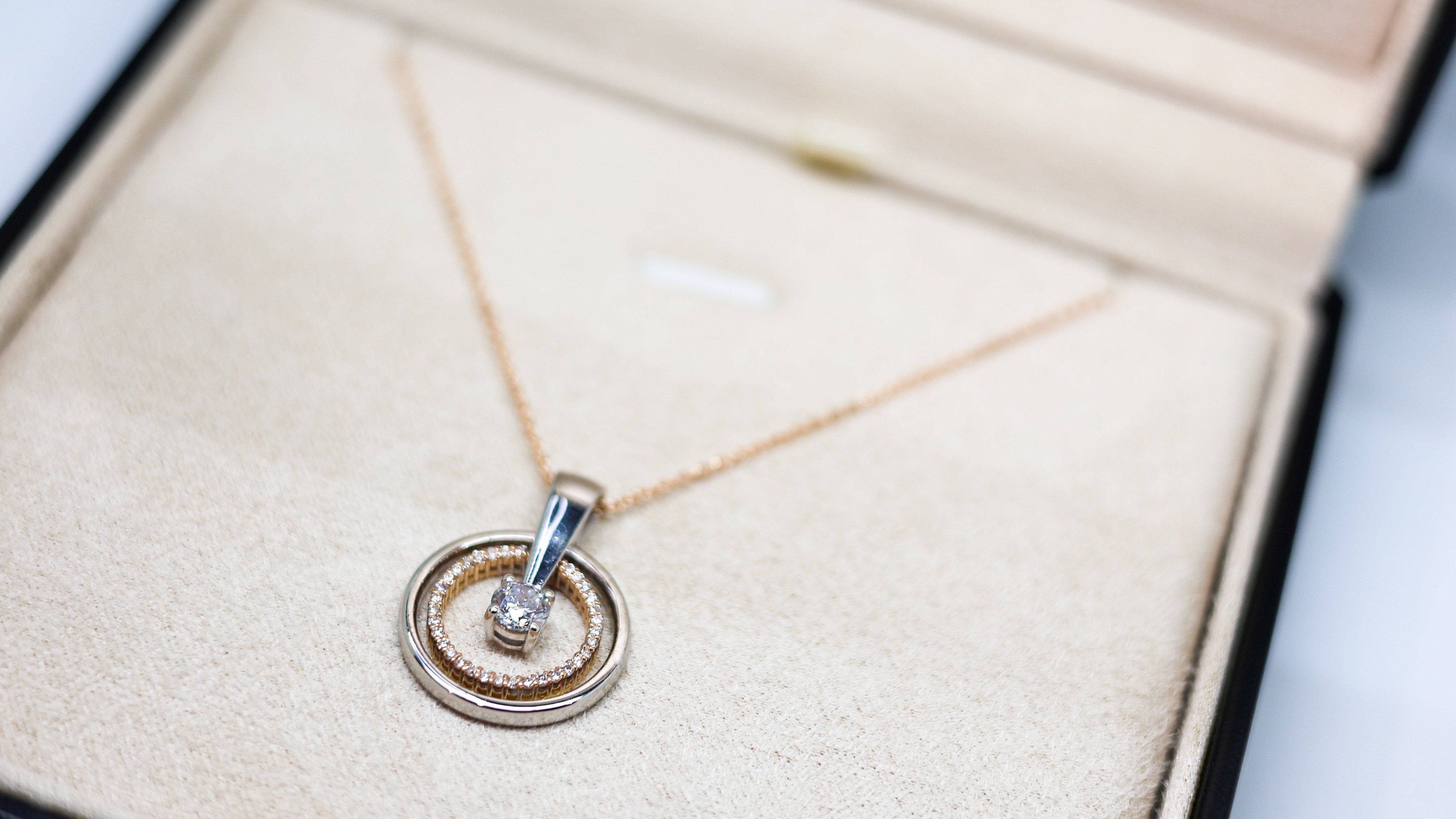 A delicate gold chain necklace with a double-ring pendant, one of which is set with a small clear gemstone, displayed inside an open beige jewelry box.