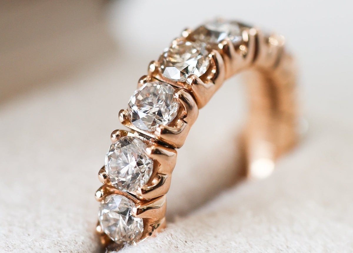 A rose gold eternity ring with five round-cut clear diamonds set in a prong setting, resting on a soft cream-colored ring box.