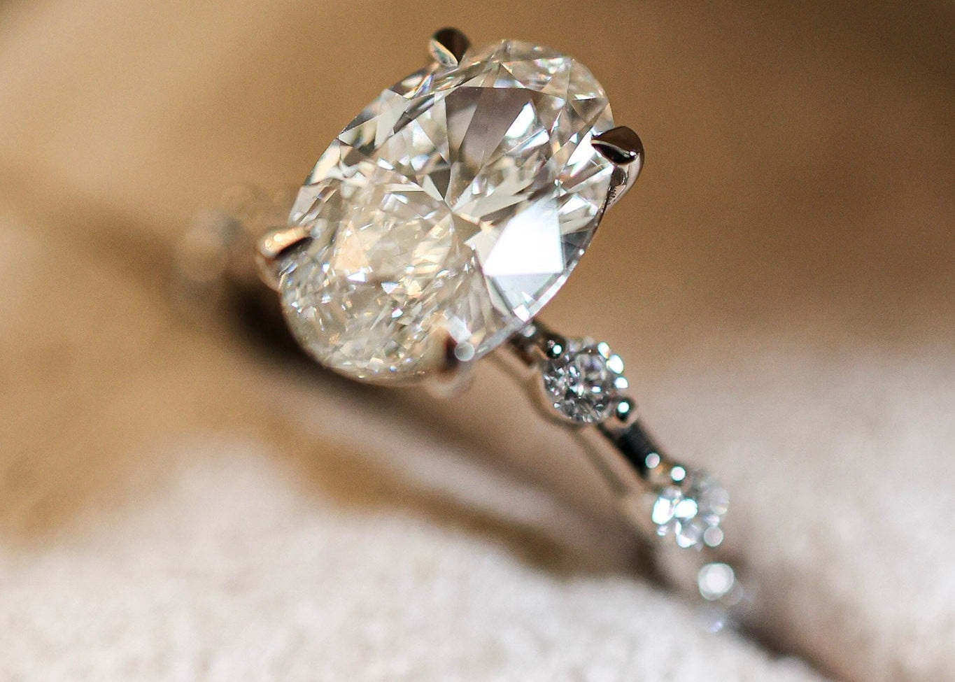 An oval-cut diamond engagement ring with a silver-toned band featuring small accent diamonds, resting on a soft beige textured surface.