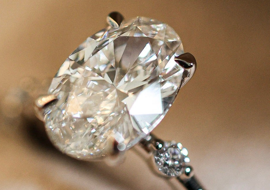 An oval-cut diamond engagement ring with a silver-toned band featuring small accent diamonds, resting on a soft beige textured surface.