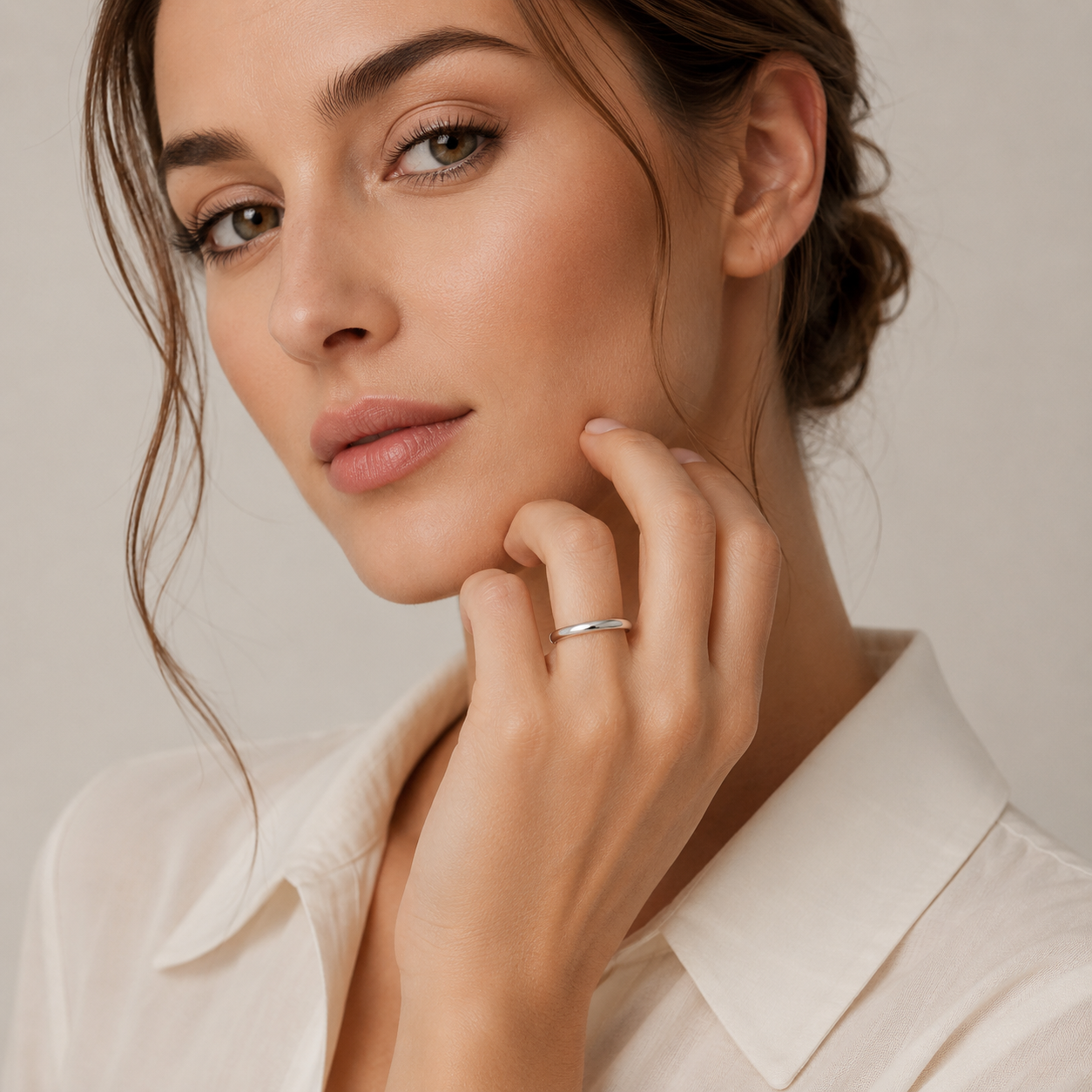 A close-up of a woman wearing a minimalist silver band ring on her finger, posing with her hand near her chin against a soft beige background.