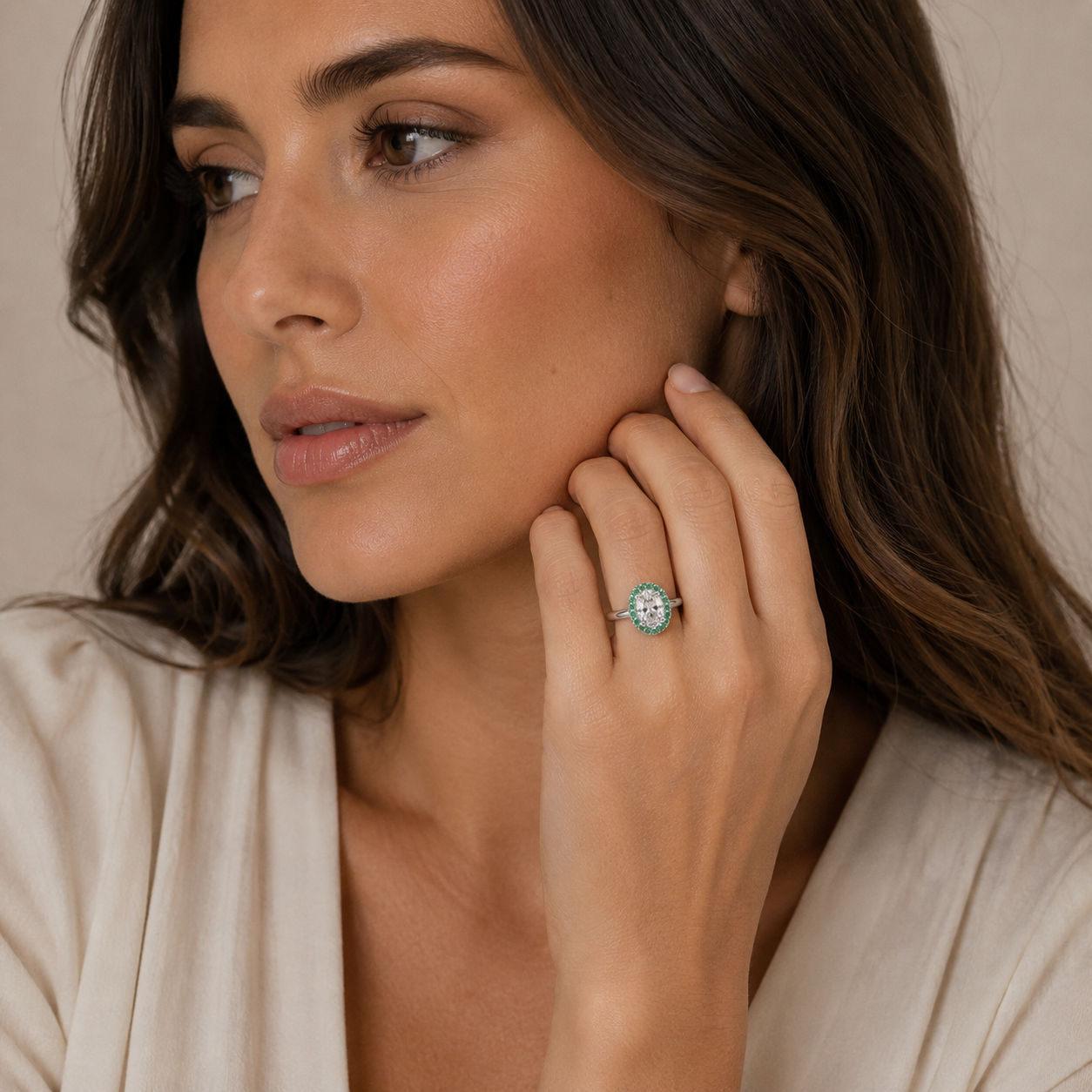 A close-up lifestyle shot of a woman wearing a diamond halo ring with a green emerald-cut center stone on her finger, set against a soft beige background.