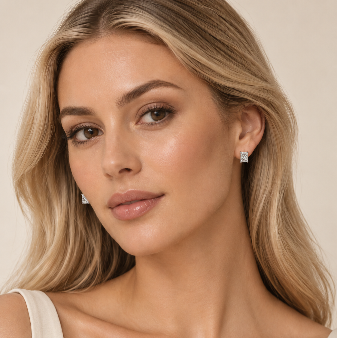 A close-up of a blonde woman wearing a pair of small square-cut silver stud earrings against a soft beige background.
