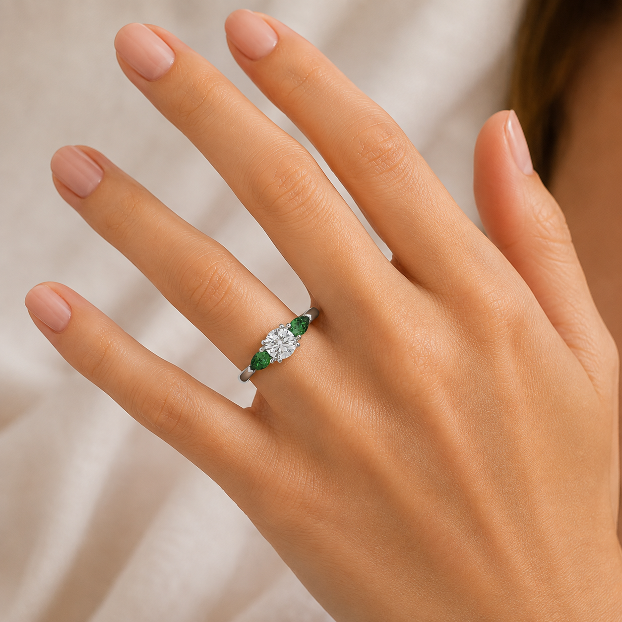 A close-up of a hand wearing a silver ring with marquise-cut green emeralds and a round brilliant-cut center stone, set against a soft beige fabric background.