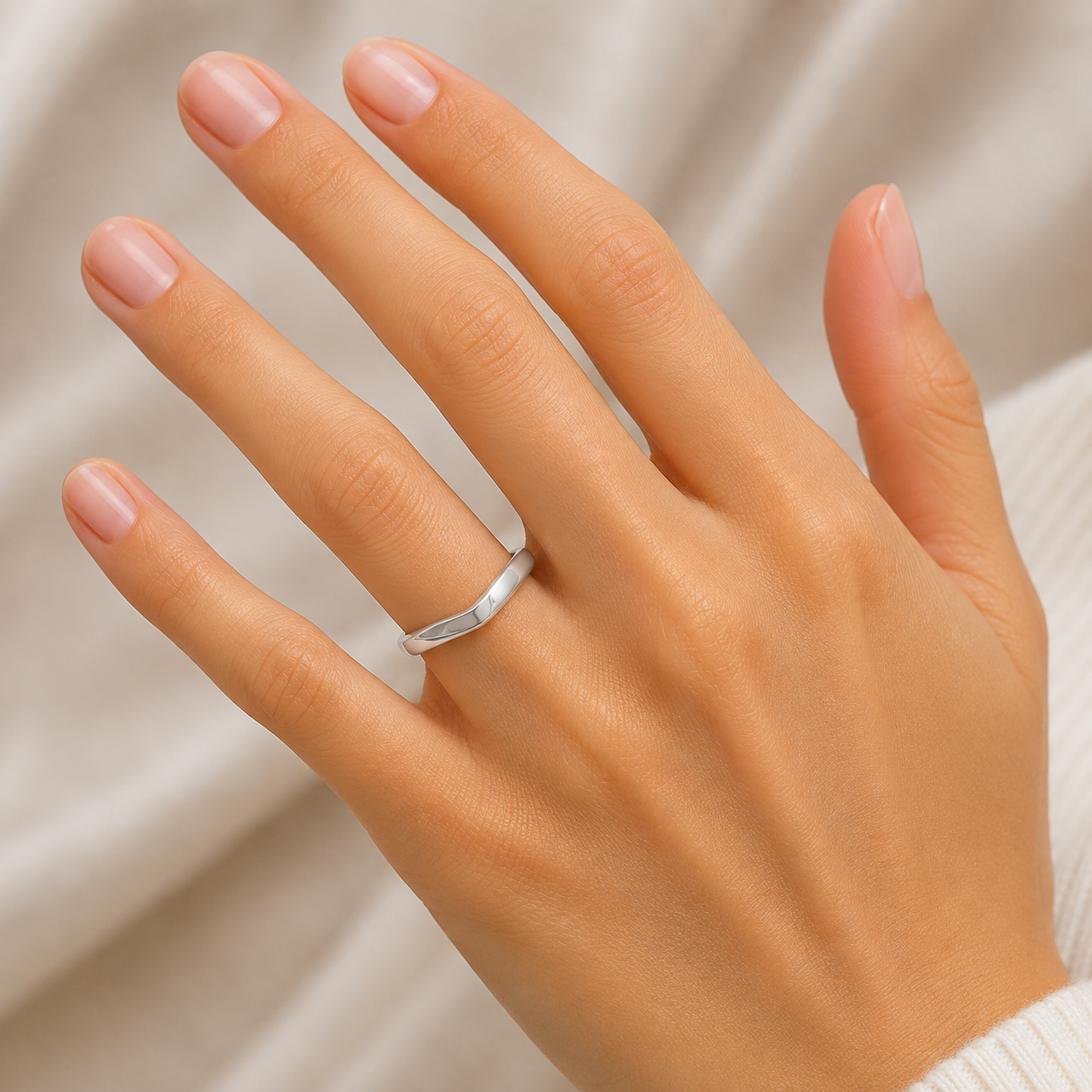 A close-up of a hand wearing a simple polished silver band ring, set against a soft beige fabric background.