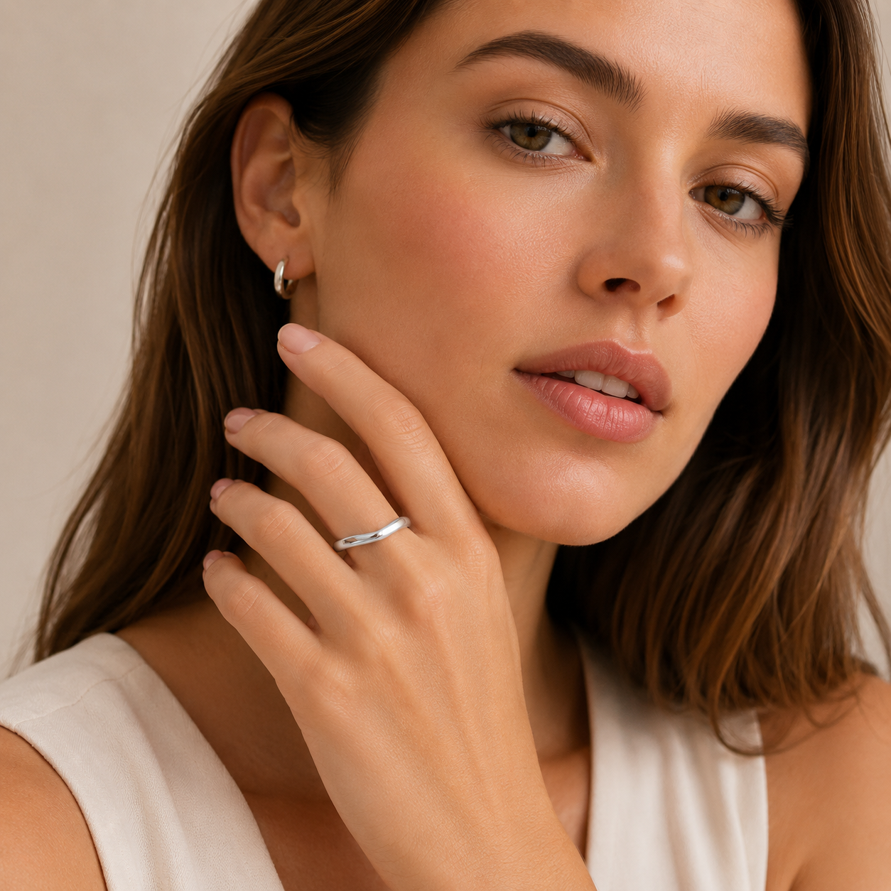 A close-up lifestyle shot of a woman wearing a thin silver band ring on her finger and a small silver hoop earring in her ear, set against a soft beige background.