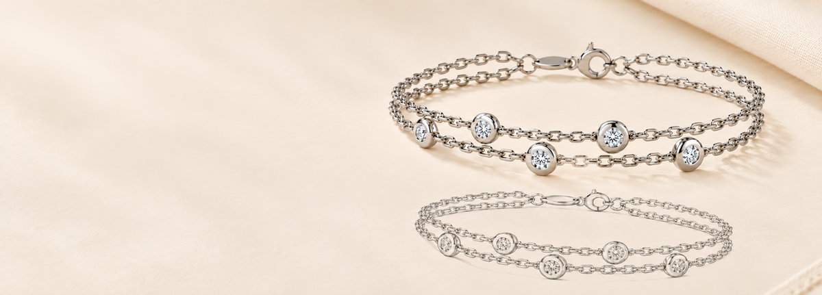 A silver-toned double-strand chain bracelet with round bezel-set clear stones, displayed on a soft beige fabric background with a mirrored reflection below.