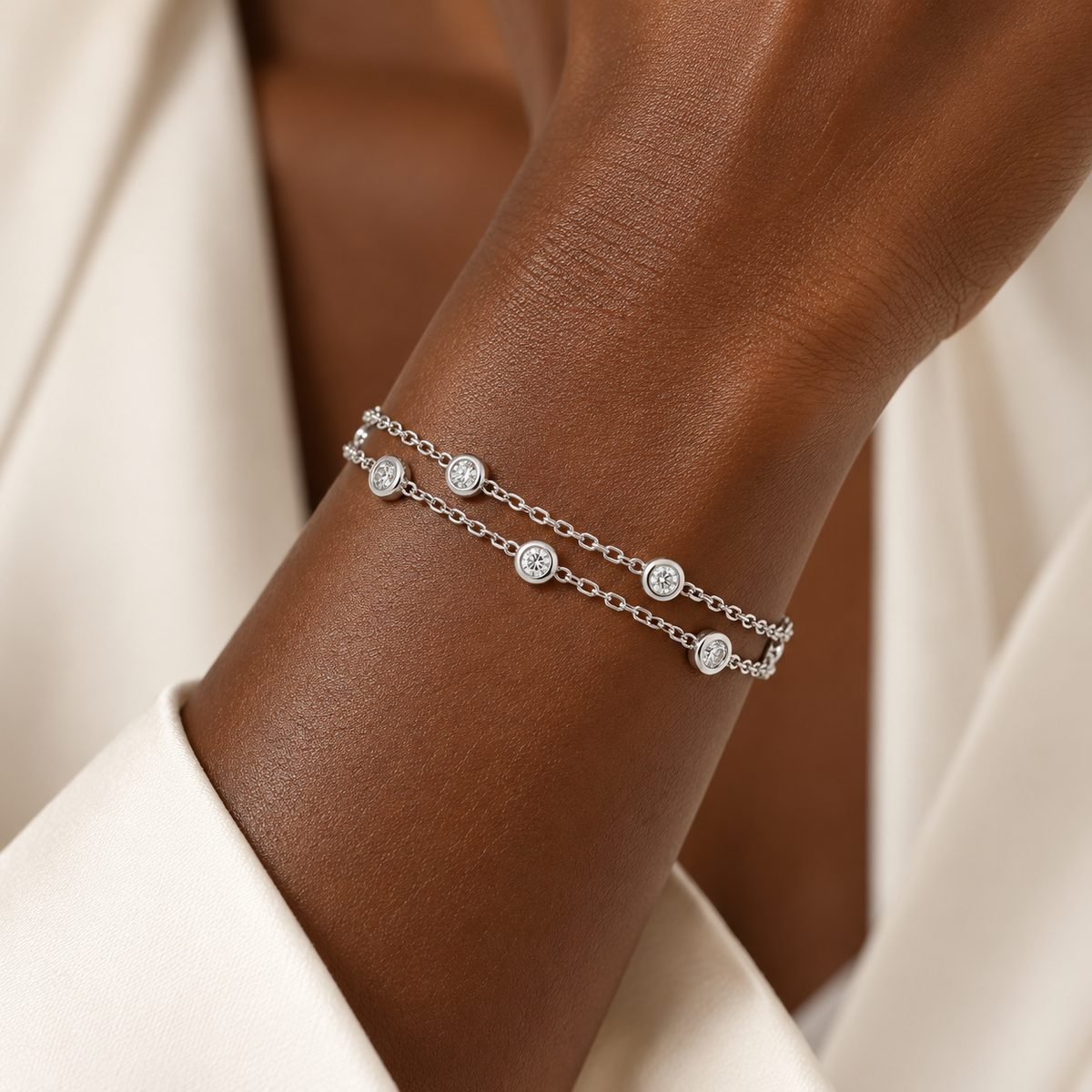A close-up of a model's wrist wearing a delicate double-strand silver chain bracelet with small round bezel-set clear stones, styled with a cream-colored top.