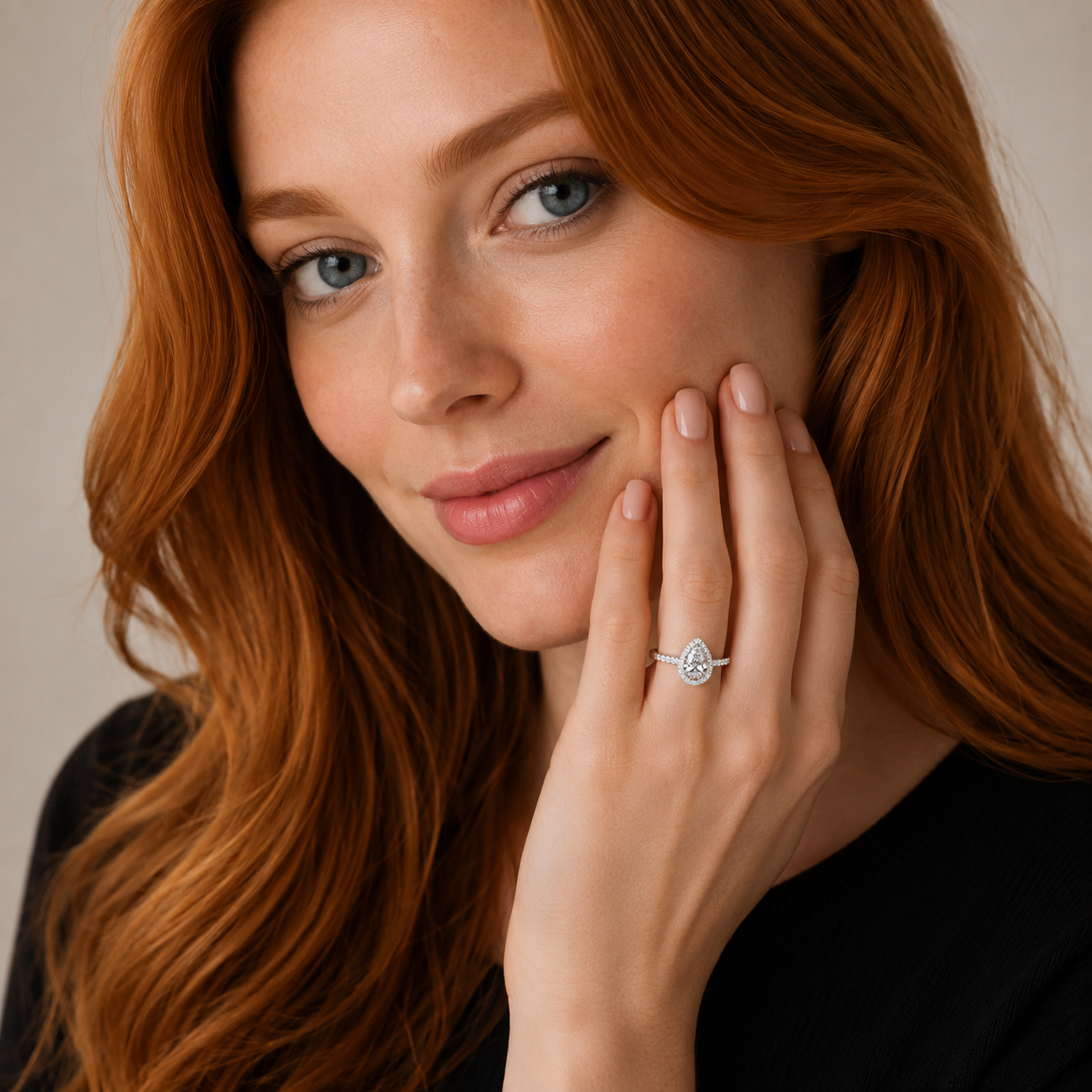 A close-up portrait of a woman with long wavy red hair wearing a silver ring with a pear-cut clear gemstone on her ring finger, set against a soft beige background.