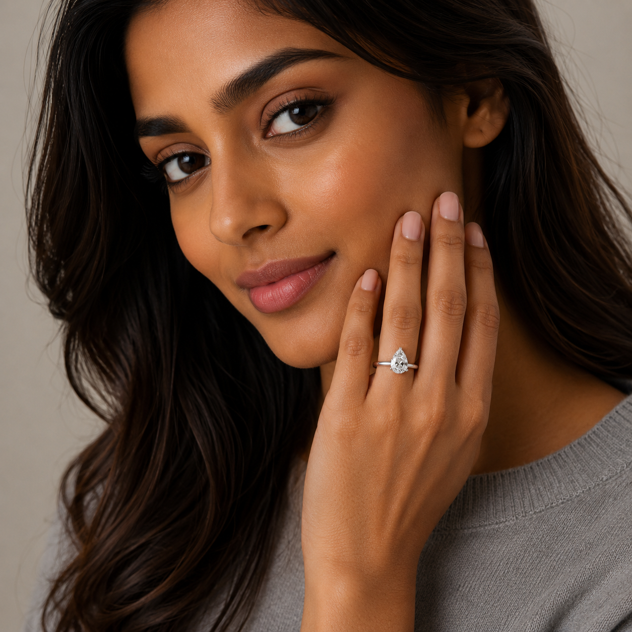 A close-up lifestyle shot of a woman wearing a diamond engagement ring on her ring finger, with her hand resting on her cheek against a soft beige background.