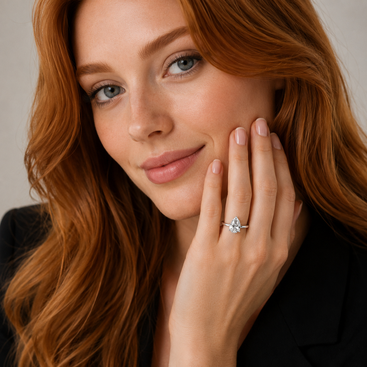 A close-up portrait of a woman with long wavy red hair wearing a delicate silver ring with a clear marquise-cut gemstone on her finger, posed against a soft neutral background.