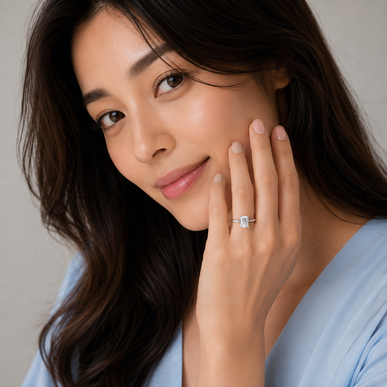 A close-up lifestyle shot of a woman with long dark brown hair wearing a delicate silver ring with a square-cut clear gemstone on her ring finger. She is wearing a light blue top against a soft, neutral background.