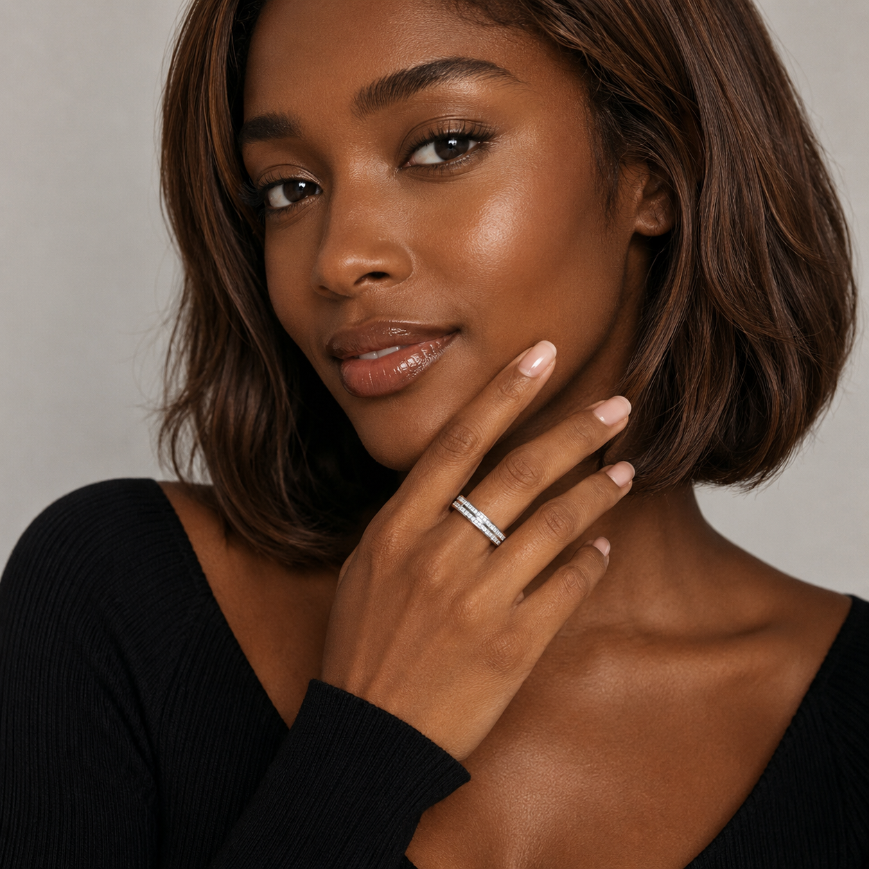 A close-up portrait of a woman with a short brown bob, wearing a silver band ring on her ring finger, against a neutral light gray background.