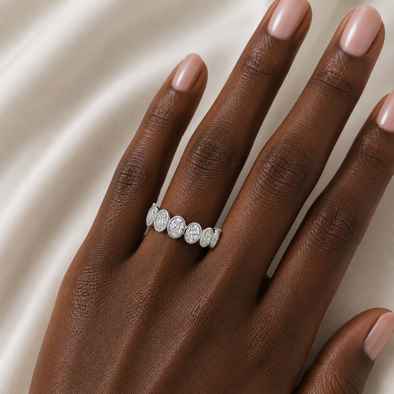 A close-up of a hand with light pink manicured nails wearing a silver oval pavé ring, resting against a soft, draped cream satin fabric background.