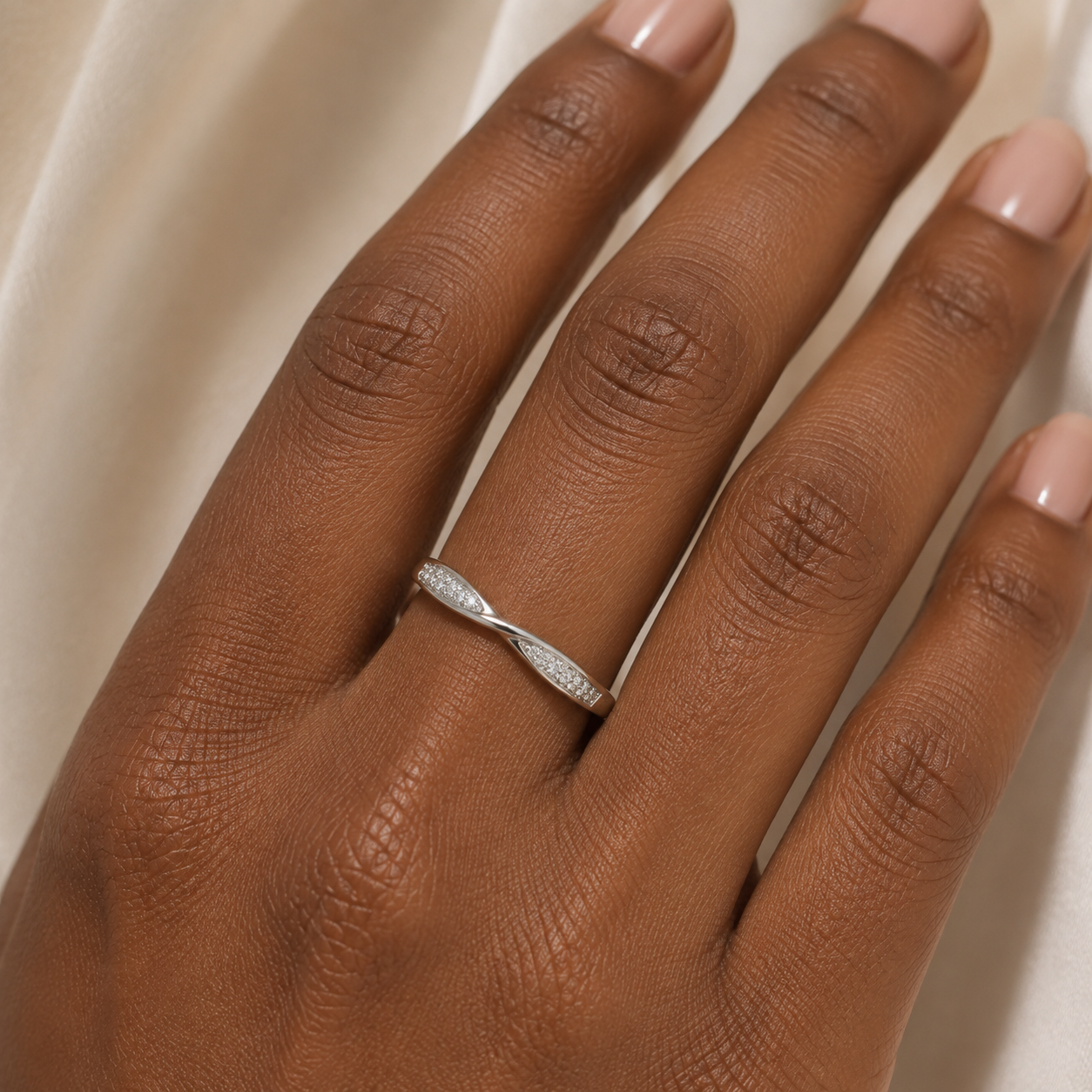 A close-up of a hand wearing a thin silver band ring with pavé-set clear stones, photographed against a soft cream fabric background.