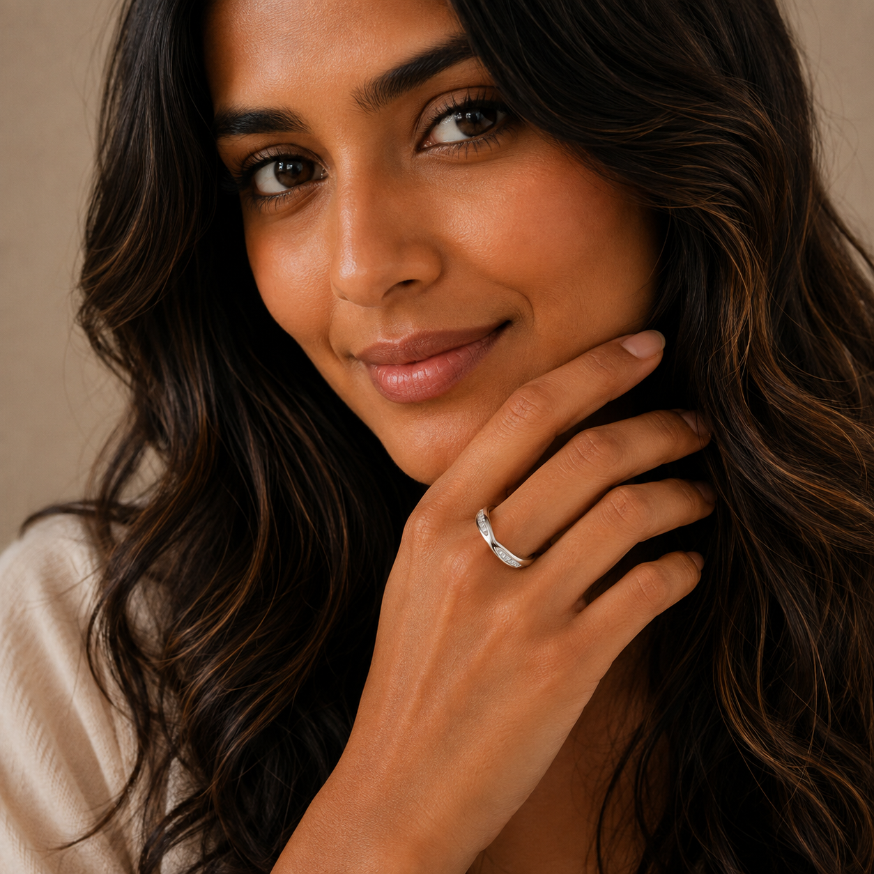A close-up lifestyle shot of a woman with long, wavy dark brown hair wearing a slim silver band ring on her finger, posing with her hand near her chin against a soft beige background.