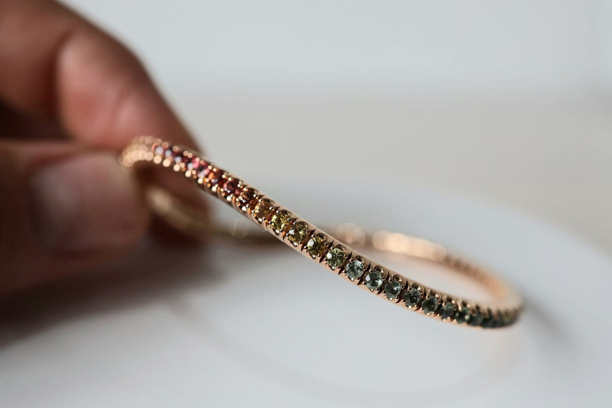 A close-up of a rose gold bangle bracelet with a row of small multicolored gemstones, held by a hand against a soft white background.