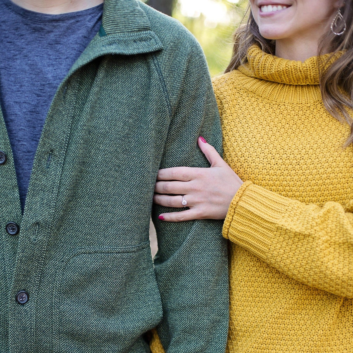 A close-up lifestyle shot of a couple holding hands outdoors. The person on the left wears an olive green button-up knit cardigan over a dark gray shirt and blue jeans.