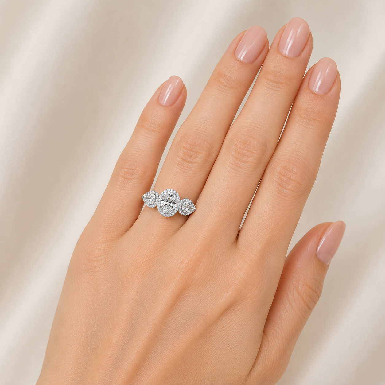 A close-up of a hand with light pink manicured nails wearing a silver ring set with three round clear gemstones, photographed against a soft beige satin fabric background.