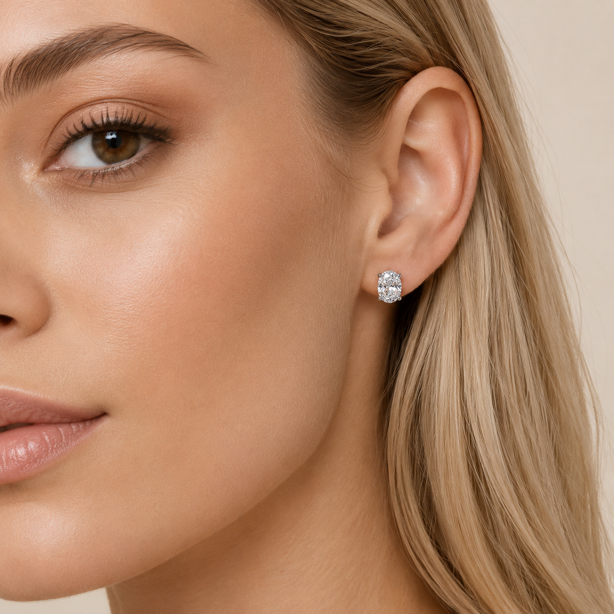 A close-up of a woman's ear wearing a small round stud earring with a sparkling clear gemstone in a silver-toned setting, against a soft beige background.