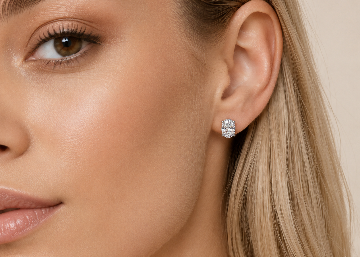 A close-up of a woman's ear wearing a small round stud earring with a sparkling clear gemstone in a silver-toned setting, against a soft beige background.