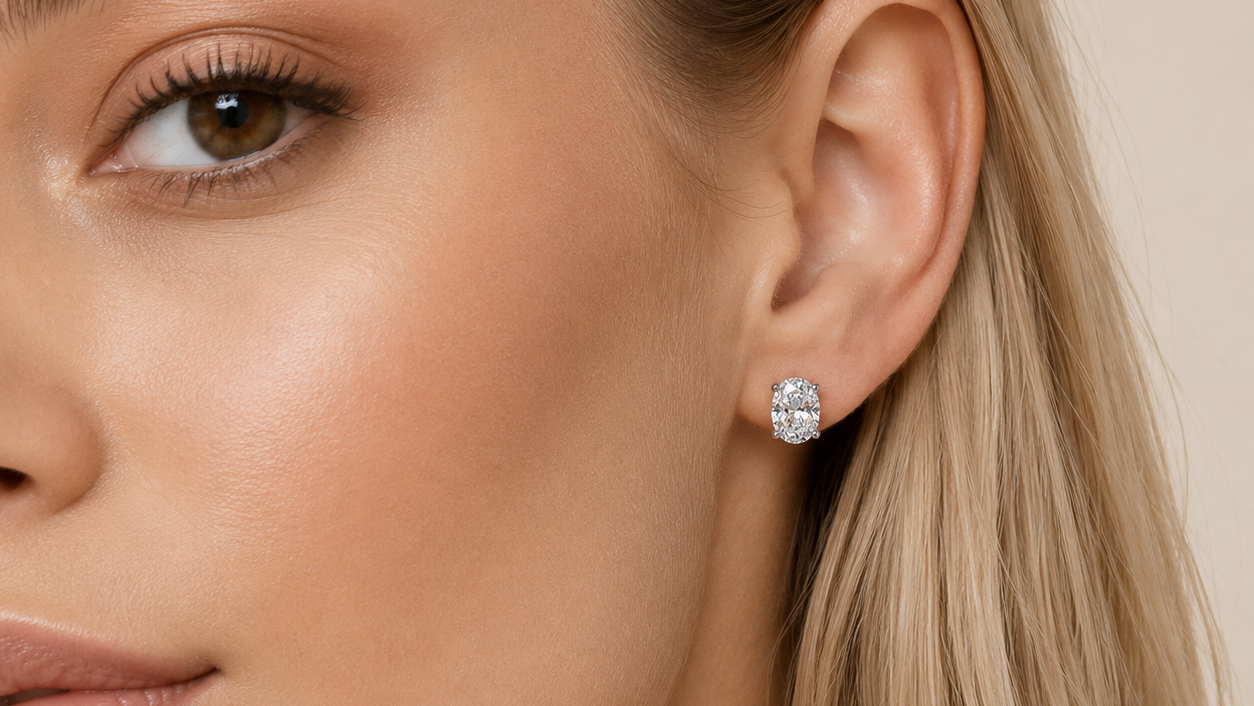 A close-up of a woman's ear wearing a small round stud earring with a sparkling clear gemstone in a silver-toned setting, against a soft beige background.