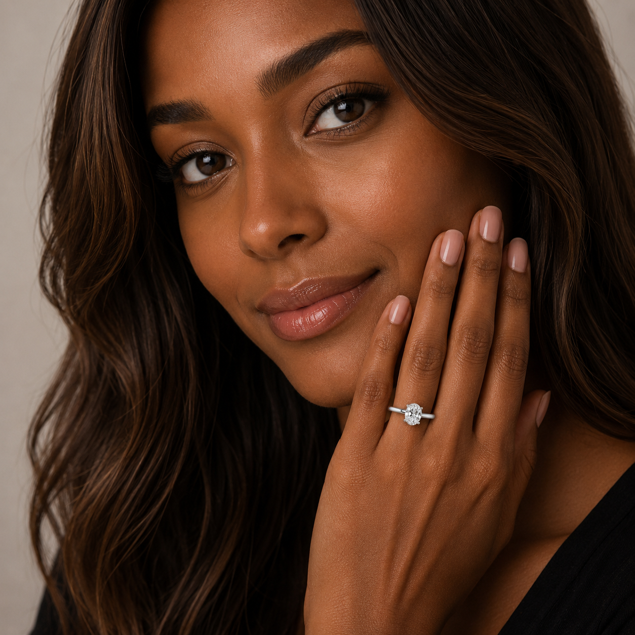 A close-up lifestyle shot of a woman with long brown hair wearing a silver engagement ring with a round-cut clear gemstone on her finger, with her hand resting against her cheek.