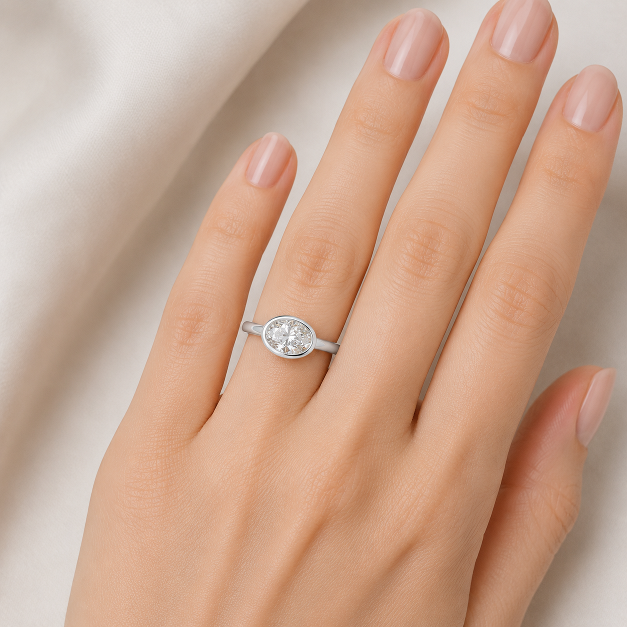 A silver oval ring with a clear gemstone set on a woman's ring finger, shown against a soft beige background.