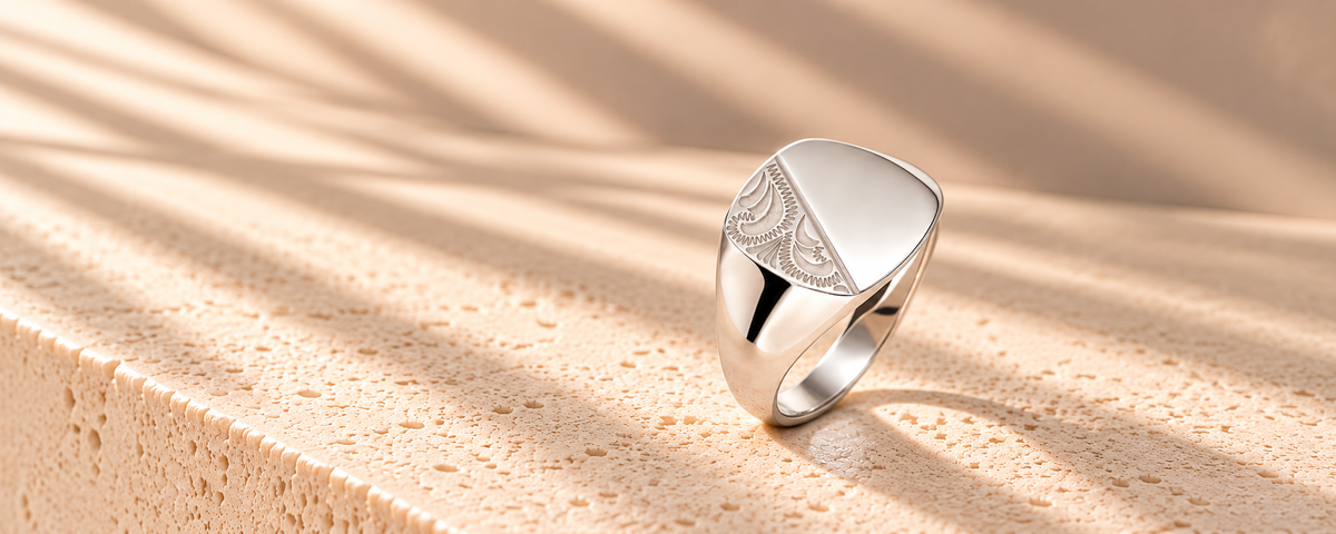 A polished silver signet ring with a shield-shaped face and engraved leaf-like detailing, resting on a textured beige stone surface under warm, dappled sunlight.