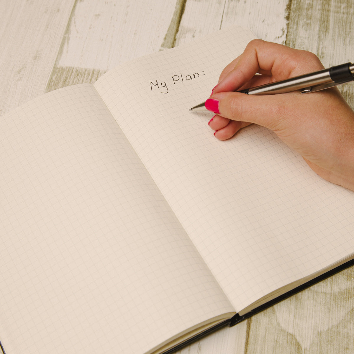 An open grid notebook on a whitewashed wooden surface, with a hand holding a pen writing in the top right corner.