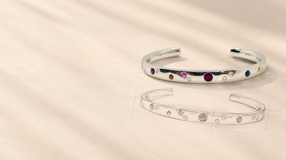 A polished silver cuff bracelet with small multicolored gemstones set along the front, displayed on a warm beige textured surface with a faint sketch of the bracelet below it.