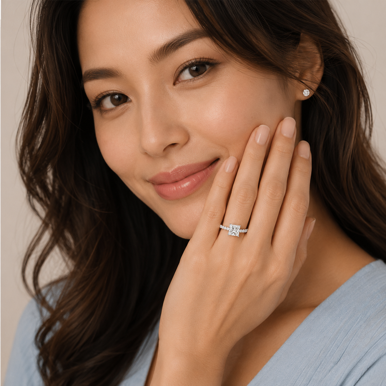 A close-up lifestyle shot of a woman with long dark brown hair, wearing a silver ring with a clear marquise-cut gemstone on her ring finger. She is wearing a small matching stud earring and a light blue ribbed top against a soft beige background.