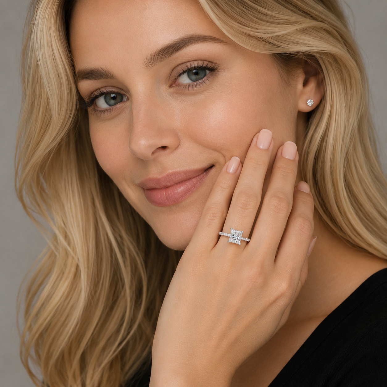 A close-up lifestyle shot of a blonde woman wearing a square-cut diamond engagement ring on her finger and a matching stud earring, set against a soft gray background.