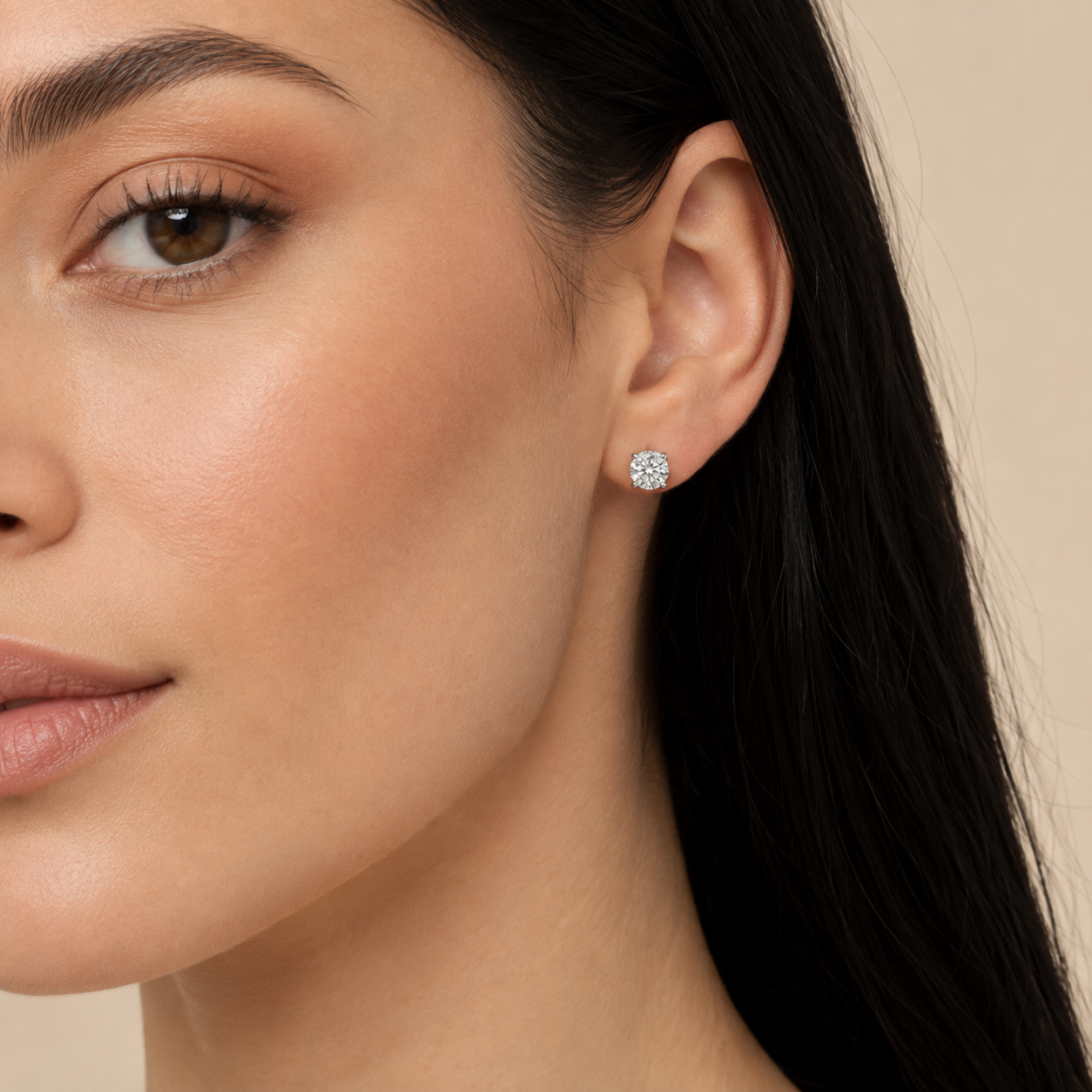 A close-up of a woman's ear wearing a small round diamond stud earring, set against a soft beige background.