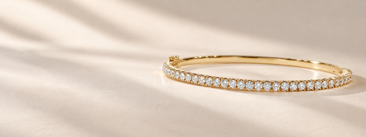 A gold tennis bracelet with a row of sparkling clear gemstones, displayed on a soft beige fabric background with elegant shadows.