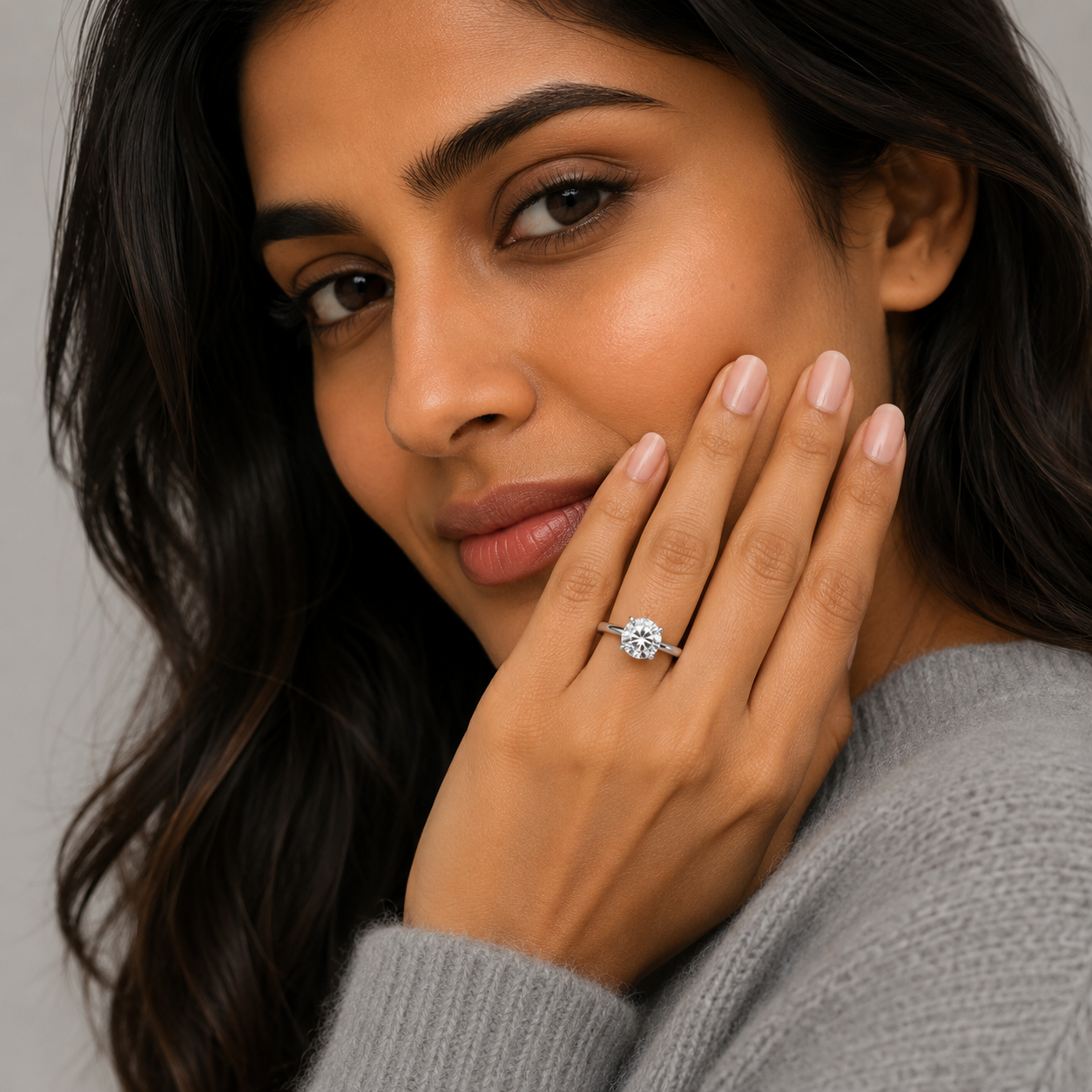 A close-up lifestyle shot of a woman wearing a round brilliant-cut diamond engagement ring on her finger, with her hand resting against her cheek. She is wearing a gray knit sweater, and the background is a soft, neutral gray.